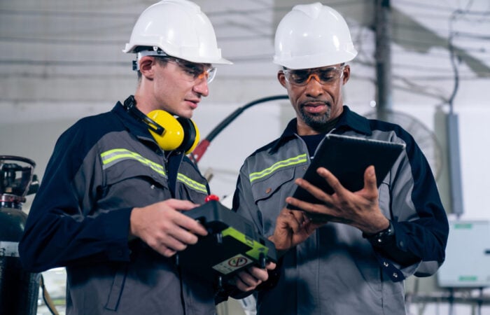Two industrial maintenance workers wearing safety helmets, goggles, and reflective uniforms are standing in a factory. One holds a diagnostic tool, while the other uses a tablet to review data or instructions. They are discussing maintenance or inspection tasks.